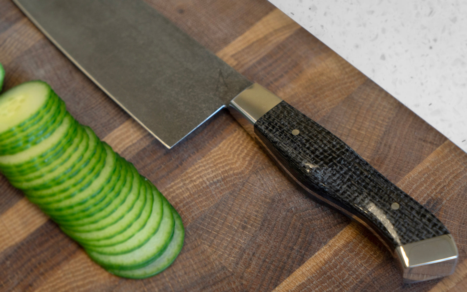 Carbon steel chef knife with black burlap handle on a wood cutting board with sliced cucumbers.